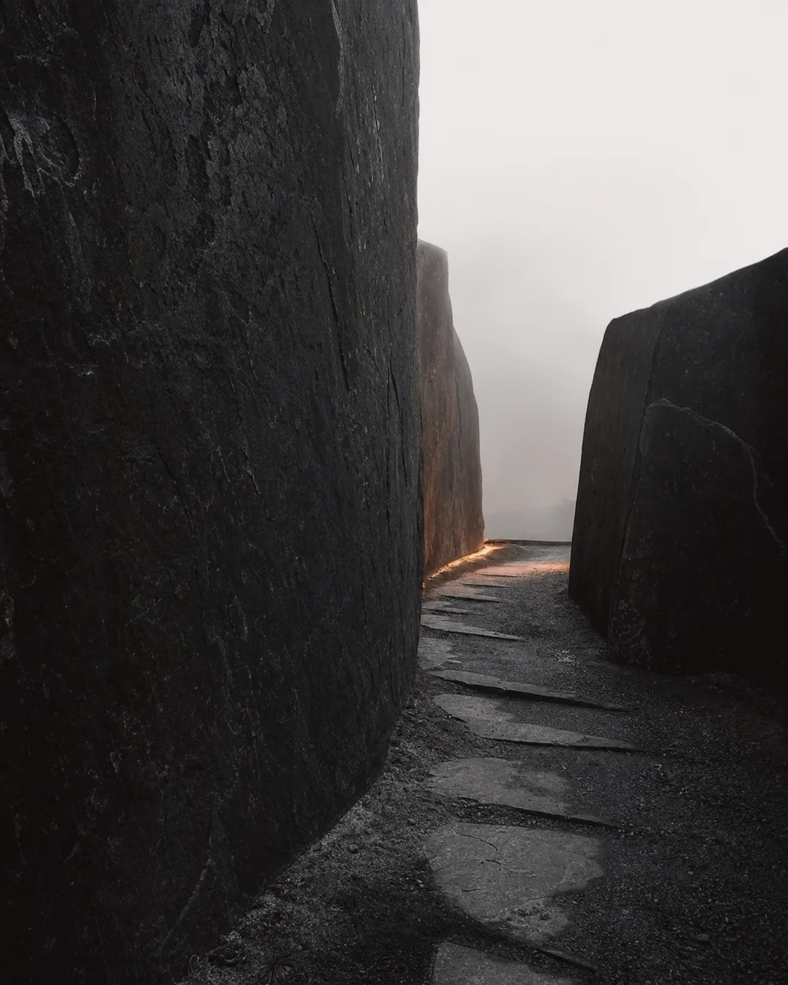 A narrow path cut between monumental dark stone planes, turning toward a restrained copper light.