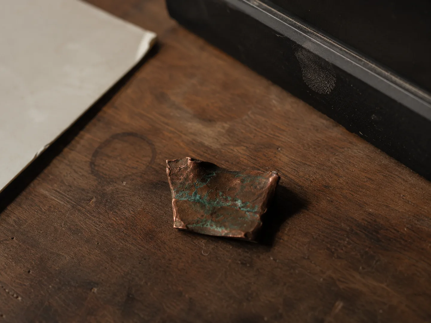 A patinated copper sample resting on a wooden desk in raking daylight.